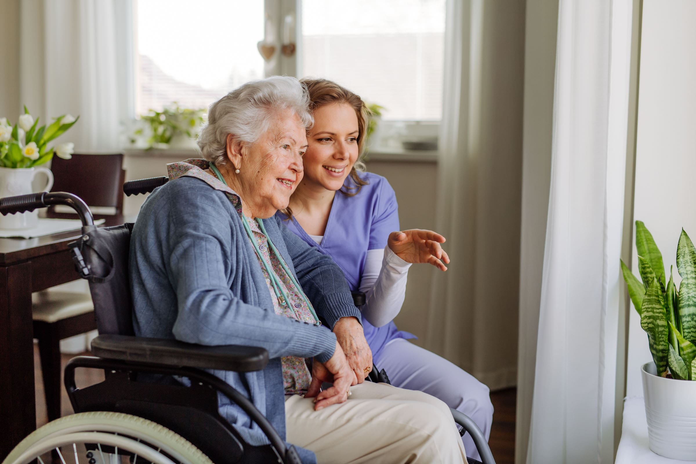 Verpleegkundige kijkt samen met oudere dame in rolstoel naar buiten